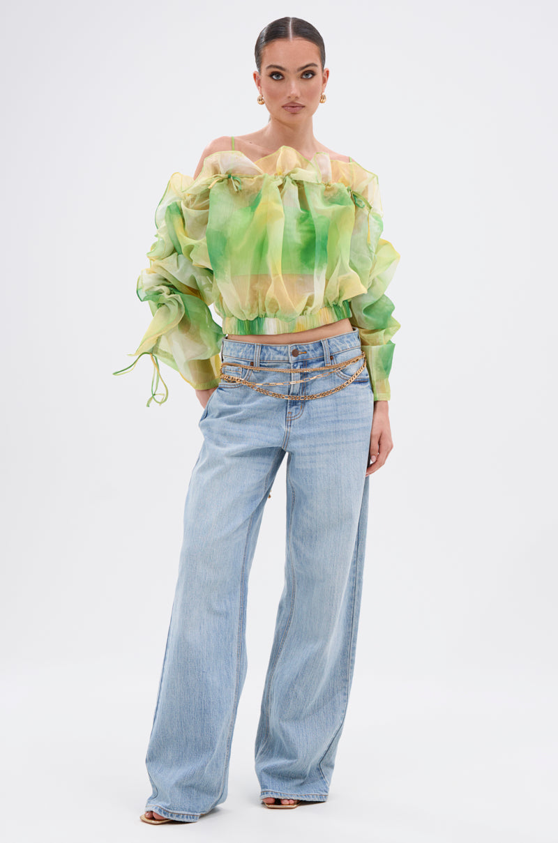 A woman poses in the LIMONE CHIFFON BLOUSE, featuring sheer green and yellow ruffles with puff sleeves, styled with light blue wide-leg jeans, a chain belt, and open-toe heels against a plain white background. 9