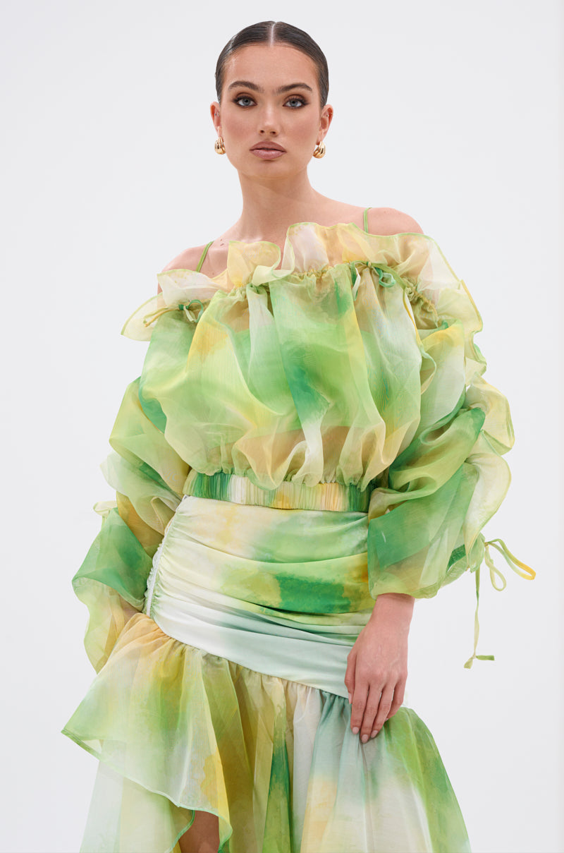 A woman poses against a plain background, wearing the LIMONE CHIFFON BLOUSE—an off-the-shoulder, flowy top with ruffled, sheer green, yellow, and white watercolor fabric. She pairs it with gold hoop earrings and has her hair slicked back. 1