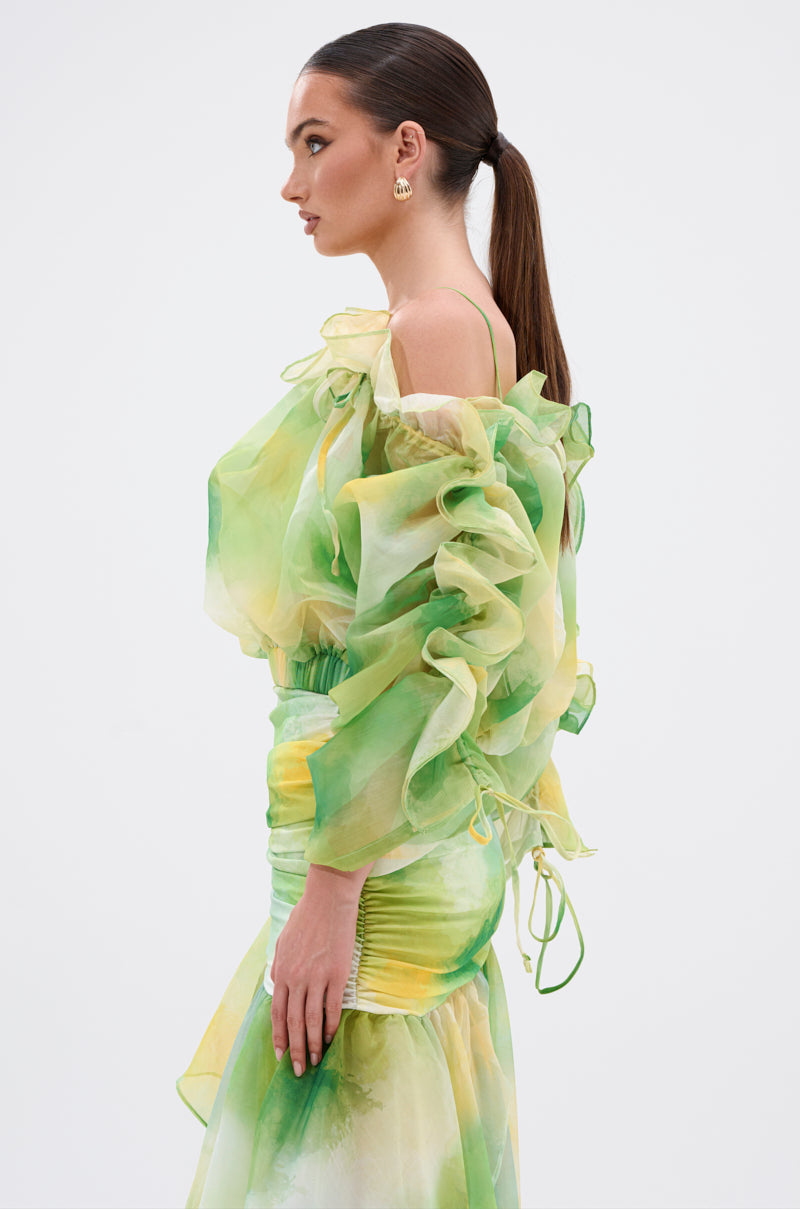 A woman with a sleek ponytail wears the LIMONE CHIFFON BLOUSE in sheer green and yellow with ruffles, ruched detailing, and an off-the-shoulder neckline, shown in profile against a plain white background. 2