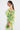 A woman with a sleek ponytail wears the LIMONE CHIFFON BLOUSE in sheer green and yellow with ruffles, ruched detailing, and an off-the-shoulder neckline, shown in profile against a plain white background.
