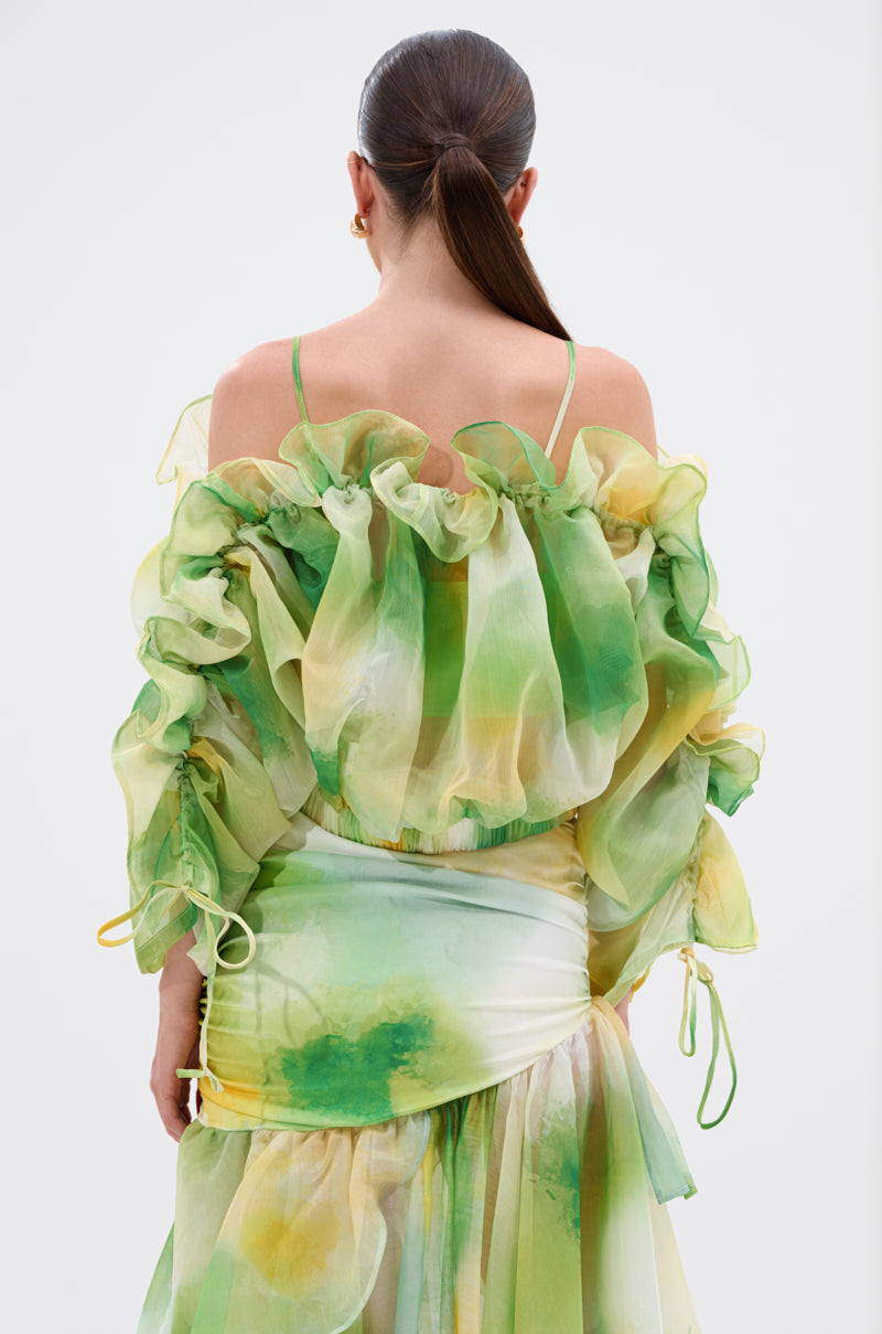 A woman with brown hair in a low ponytail is seen from behind against a plain background, wearing the LIMONE CHIFFON BLOUSE—an off-the-shoulder top with ruffled details and green, yellow, and white watercolor patterns. 3