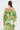 A woman with brown hair in a low ponytail is seen from behind against a plain background, wearing the LIMONE CHIFFON BLOUSE—an off-the-shoulder top with ruffled details and green, yellow, and white watercolor patterns.
