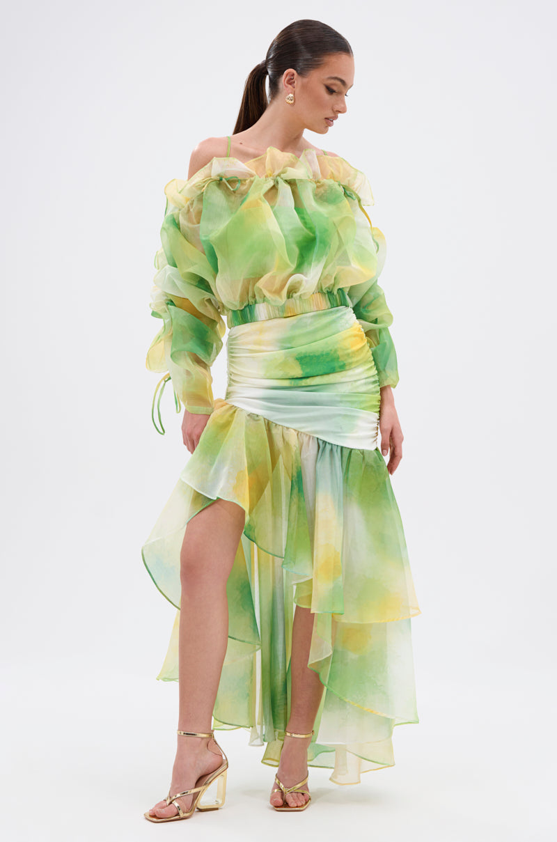 A woman styles the LIMONE CHIFFON BLOUSE, featuring an off-the-shoulder neckline, sheer puff sleeves, and a green-yellow-white watercolor print. Its ruched waist and flowy design pair perfectly with gold heels and her chic ponytail. 4