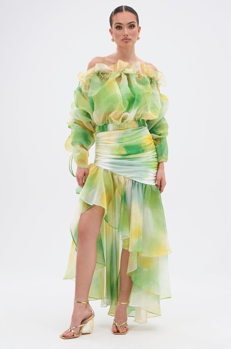 A woman poses in the LIMONE CHIFFON BLOUSE, featuring a light green-yellow watercolor style with sheer long sleeves and a ruffled neckline. She pairs it with gold strappy heels and wears her hair in a sleek bun. 5