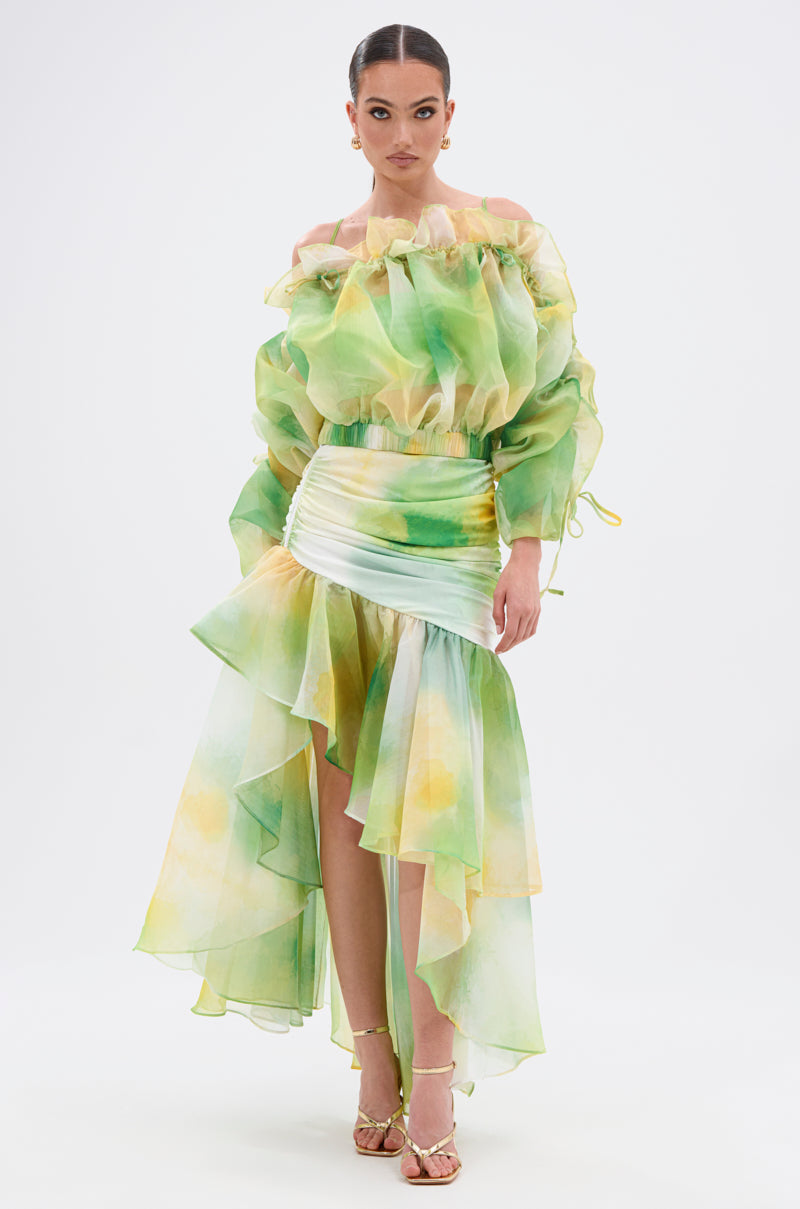 A woman wears the LIMONE CHIFFON BLOUSE with ruffled, semi-sheer green, yellow, and white fabric. The blouse features puff sleeves and an off-the-shoulder neckline. She stands in strappy heels against a white background. 6