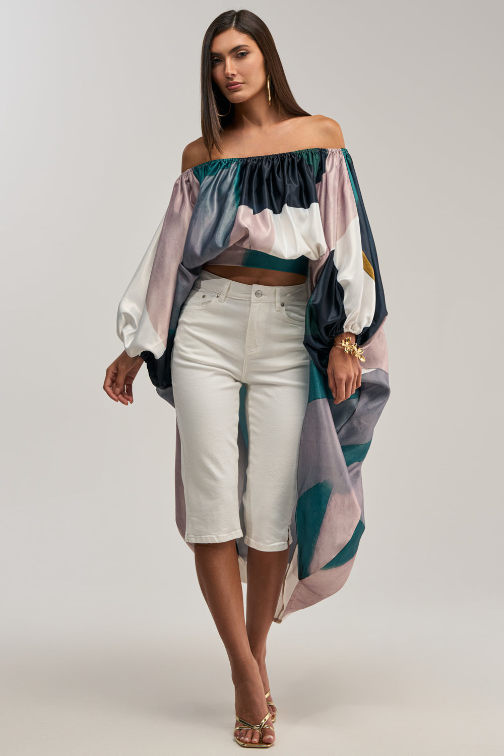 A woman wears the BE WITH ME BLOUSE IN GREEN MULTI, featuring an off-the-shoulder design, loose long sleeves, and an asymmetrical high-low hem, paired with white knee-length shorts and gold heels. She has straight brown hair and a neutral expression.