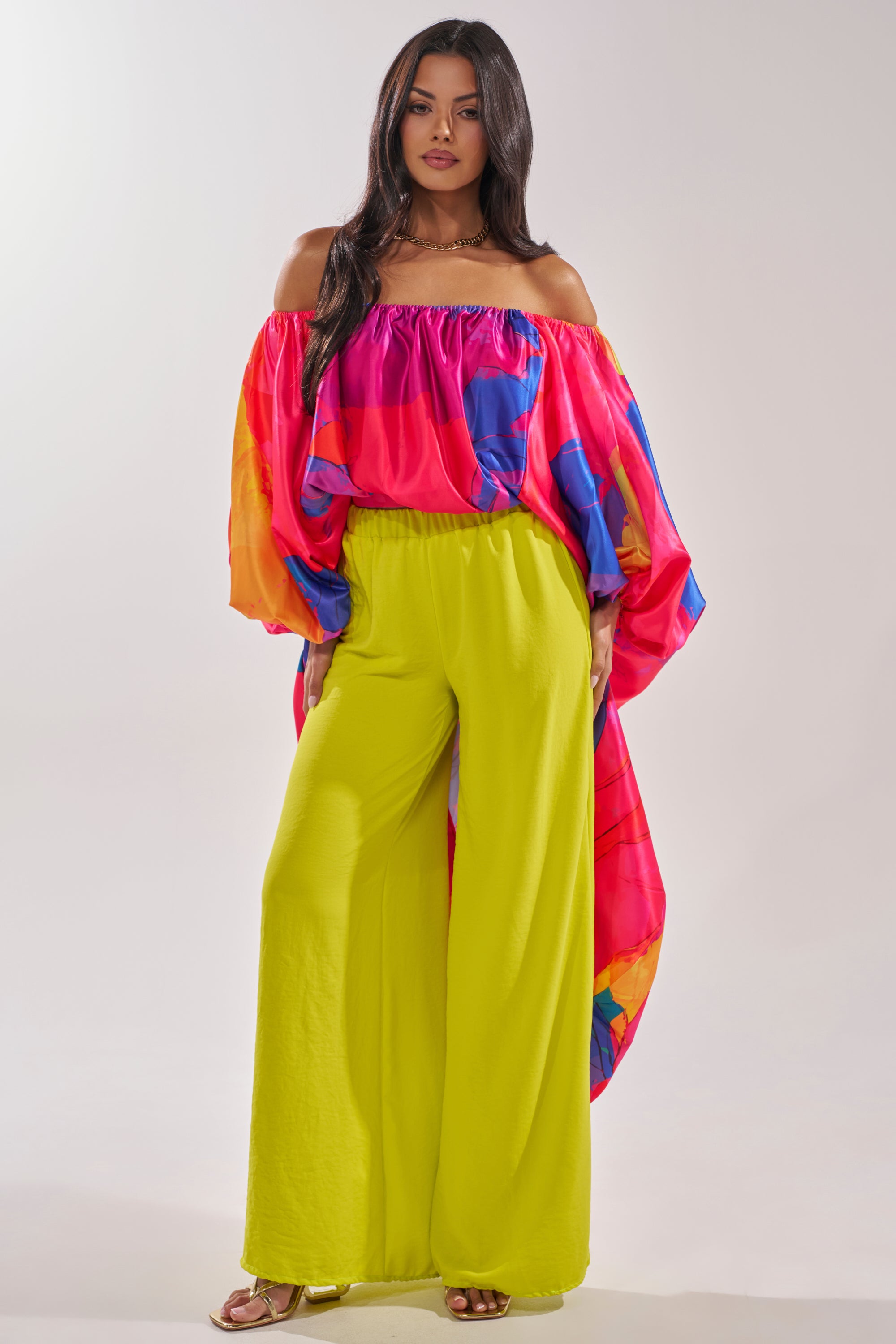 A woman with long dark hair wears the BE WITH ME BLOUSE—bright, high-low style with pink, blue, and orange patterns—and wide-legged neon yellow pants. She completes her look with gold sandals, perfect for a stylish vacation. 1
