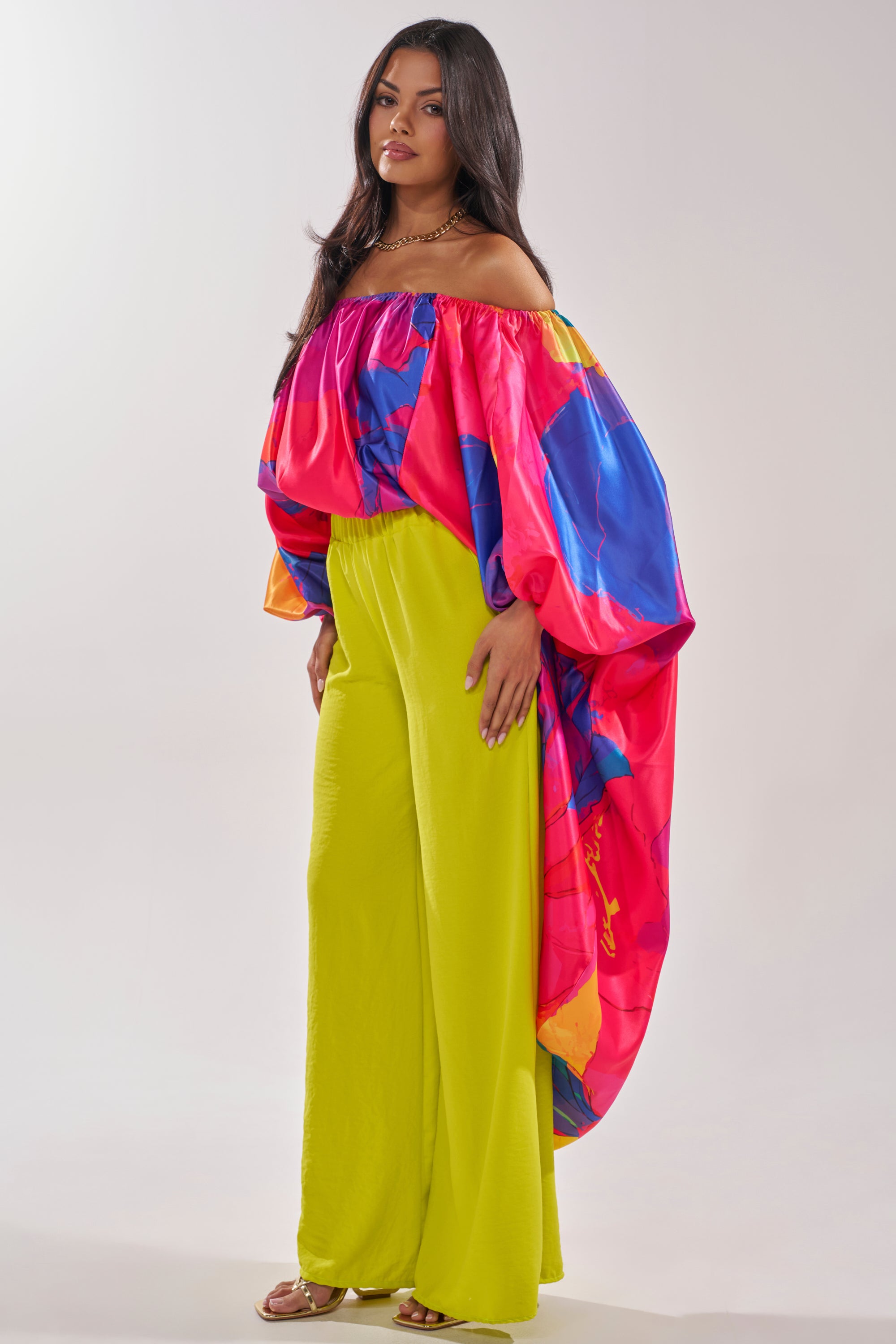 A woman wears the BE WITH ME BLOUSE, an off-the-shoulder pink top with bold blue and orange prints. She pairs it with bright yellow wide-leg pants and sandals, completing her vibrant vacation look. 3