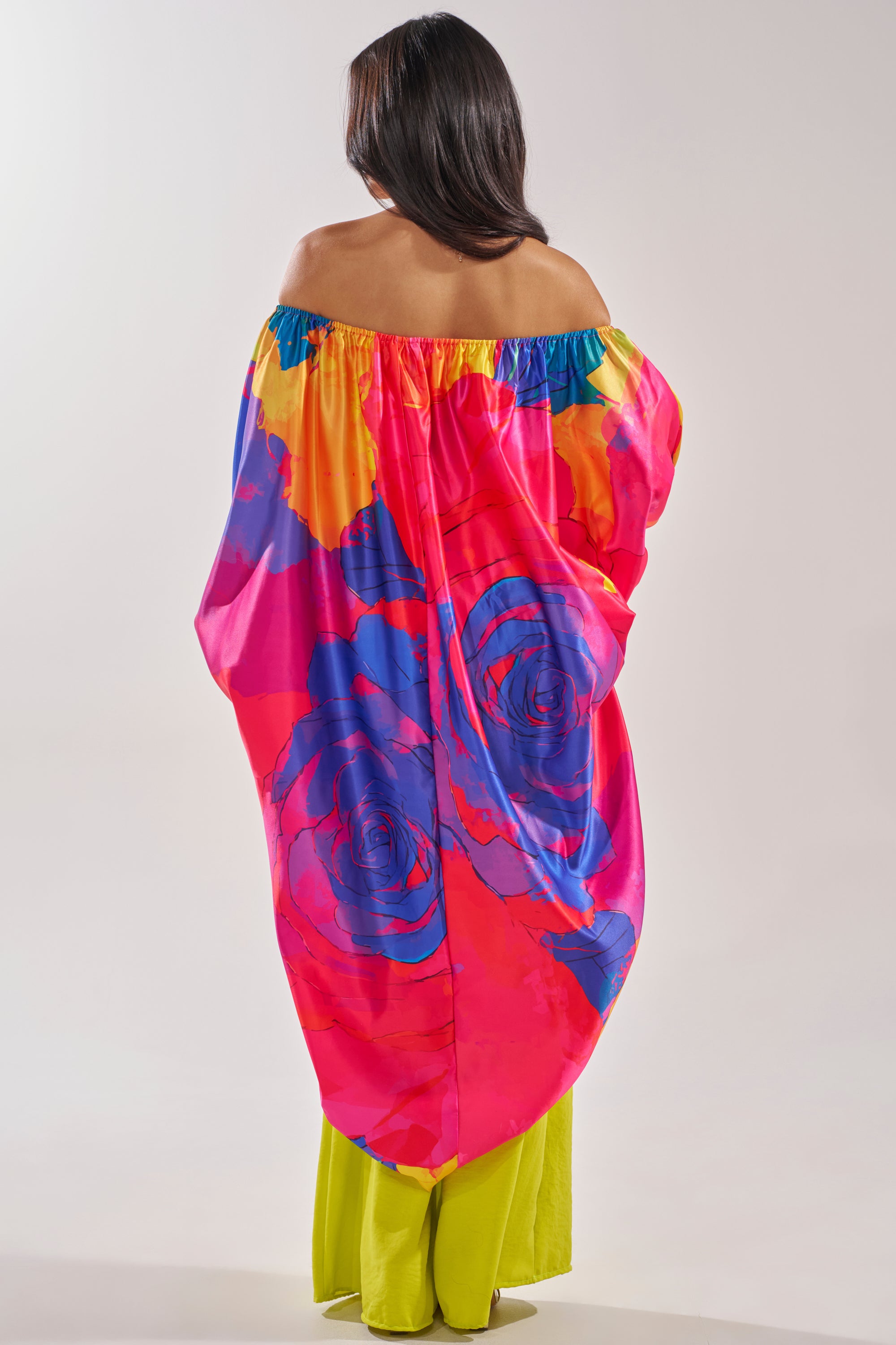 A woman with long dark hair faces away, wearing the BE WITH ME BLOUSE—an off-the-shoulder top featuring bold pink, blue, yellow, and orange abstract florals over bright yellow pants—a standout for stylish vacation outfits. 4