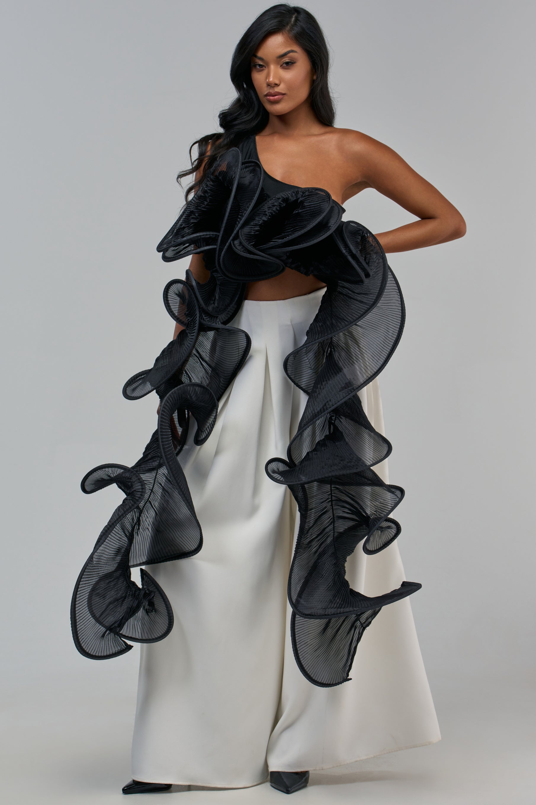 A model wears the DRAMA RUFFLE BLOUSE IN BLACK, a one-shoulder cropped blouse with large, structured circular ruffles, paired with high-waisted wide-leg white trousers.