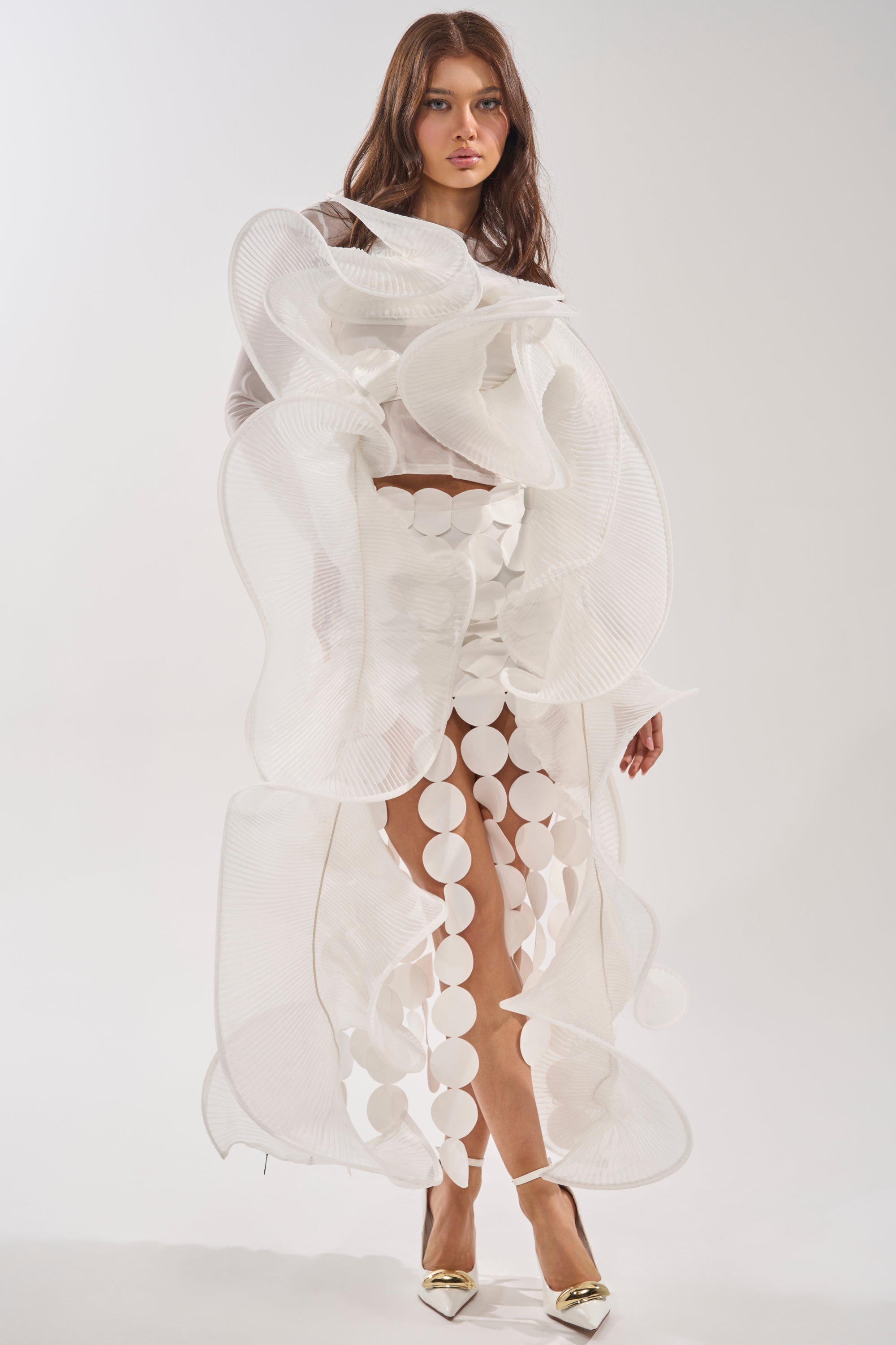 A woman poses in the DRAMA RUFFLE BLOUSE, a white avant-garde top with dramatic ruffles and large circular cutouts, styled with gold pointed-toe heels against a plain light background. 0