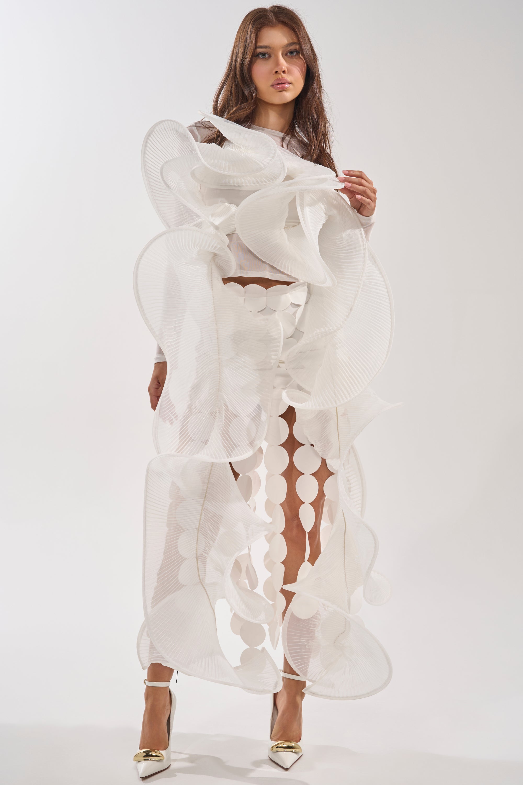 A woman stands against a plain white background wearing the DRAMA RUFFLE BLOUSE—an avant-garde white top with sculptural, wavy, sheer ruffles and circular accents—styled with metallic pointed-toe heels. 1