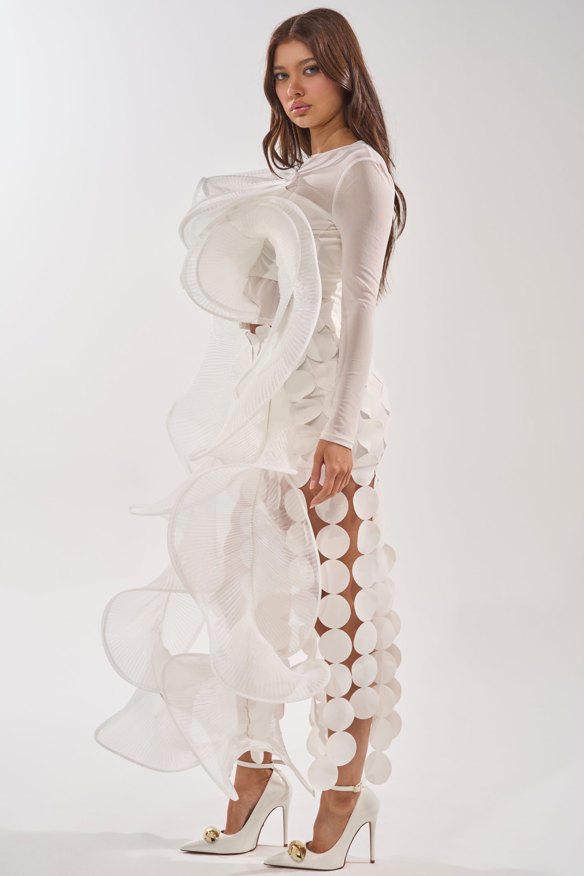 A woman wears the DRAMA RUFFLE BLOUSE, a white avant-garde long sleeve top with large sculptural ruffles and circular cutouts, styled with white high heels. She poses against a plain white background. 2