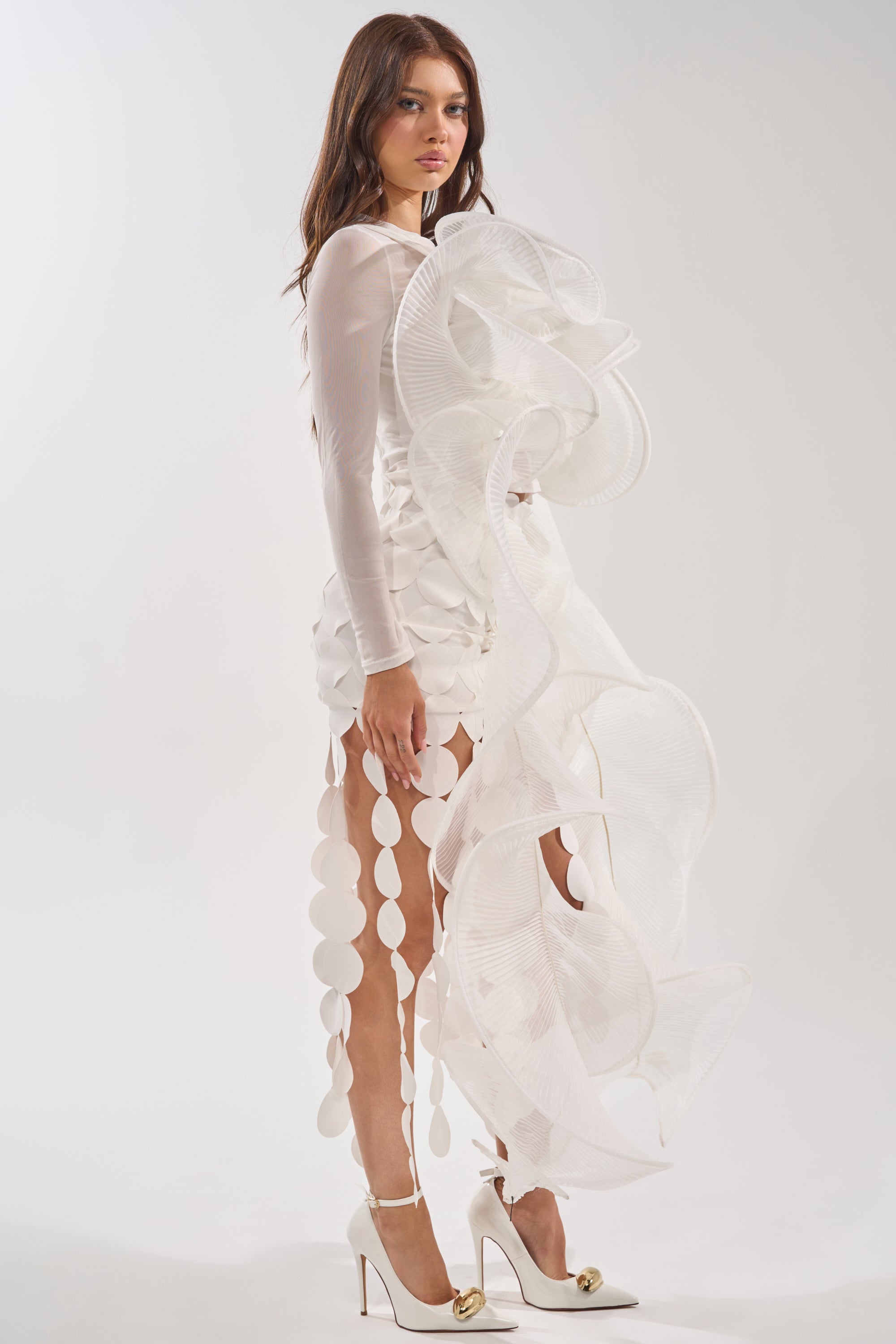 A woman in the DRAMA RUFFLE BLOUSE, featuring avant-garde white sheer sleeves and bold ruffled circular details, pairs it with high heels with gold accents, standing against a plain light background. 3