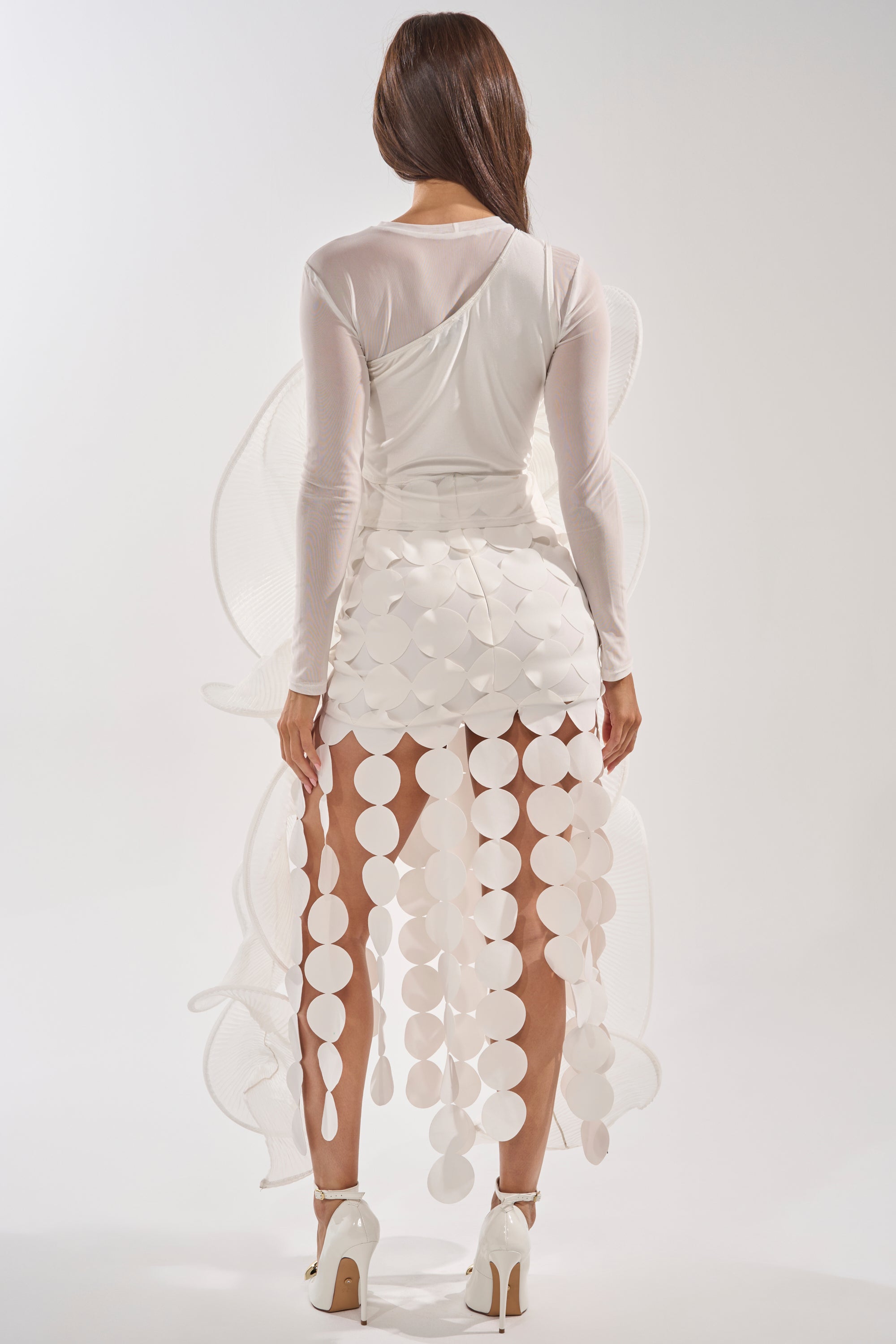 A woman faces away, wearing the DRAMA RUFFLE BLOUSE—a sheer white top with ruffled long sleeves and a fitted bodice—paired with a skirt featuring circular cutouts, white high heels, and her long brown hair cascading down her back. 4
