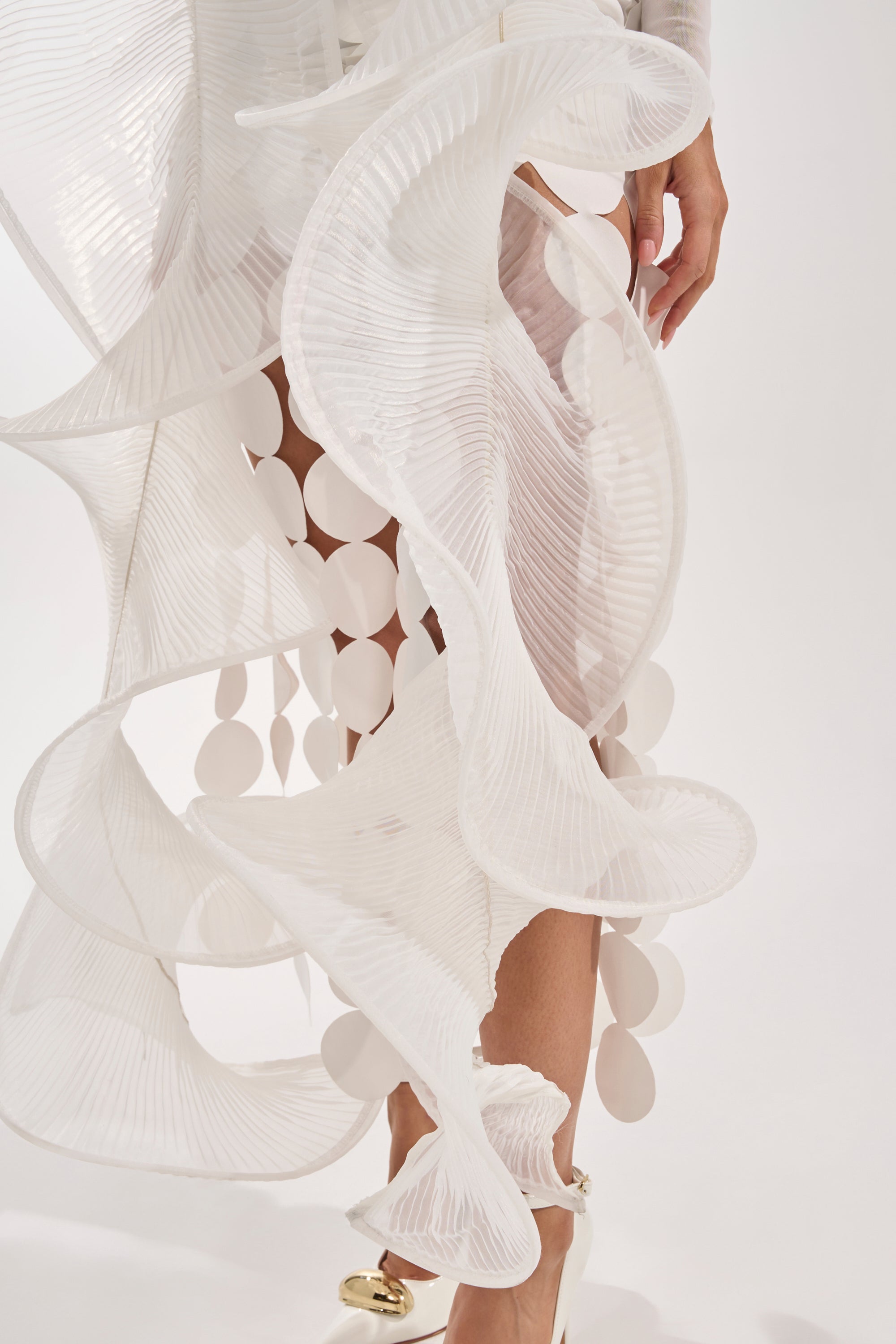 A person wears the DRAMA RUFFLE BLOUSE, an avant-garde, sculptural white top with pleated, wavy textures and dangling oval accents, styled with white high-heeled shoes against a plain light background. 6
