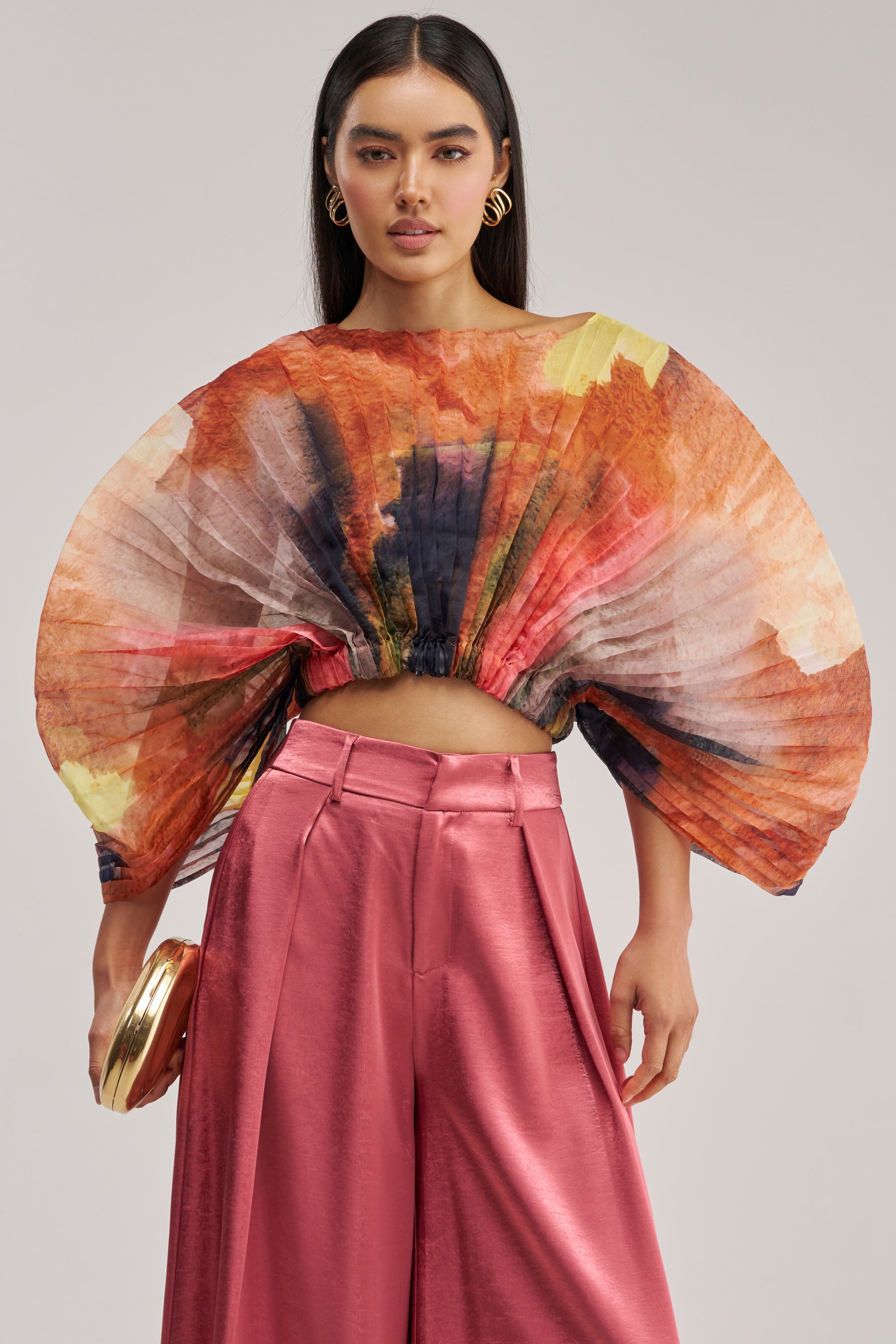 A woman wears the WATERCOLORS CHIFFON FAN TOP, a wide pleated, multicolored chiffon piece with dramatic sleeves and a playful cropped silhouette. She pairs it with high-waisted shiny pink pants, accessorizing her look with gold hoop earrings and a gold clutch. Her long dark hair is worn down and she has a neutral expression.