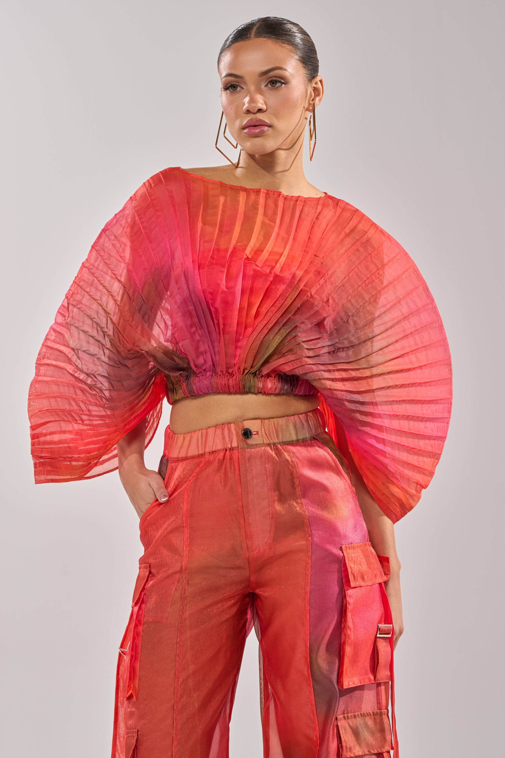 A woman wears the STAINED RED CROPPED BLOUSE with dramatic, fan-like sleeves and matching high-waisted cargo pants, standing with one hand in her pocket against a plain background. 0