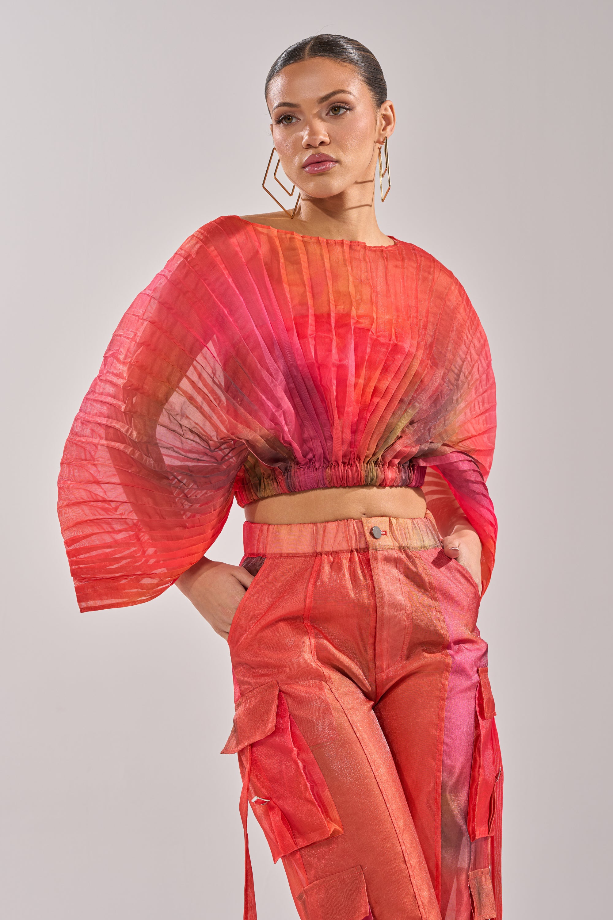 A woman models the STAINED RED CROPPED BLOUSE, a sheer chiffon fan-top with wide sleeves and cinched waist, paired with high-waisted shiny pants with decorative straps and large geometric earrings. 1