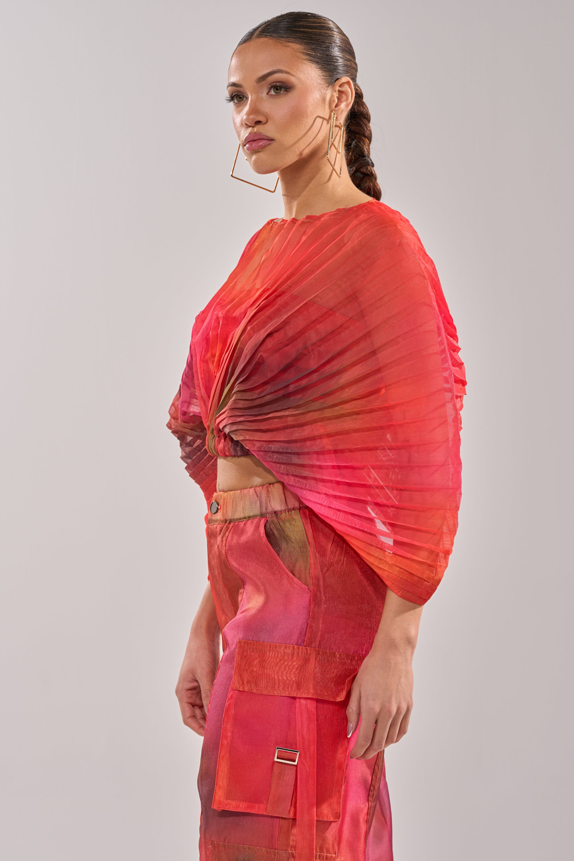 A woman models the STAINED RED CROPPED BLOUSE, featuring pleats in vibrant red and orange, paired with matching high-waisted pants. Her hair is slicked back in a braid, accessorized with large geometric hoop earrings against a plain backdrop. 2