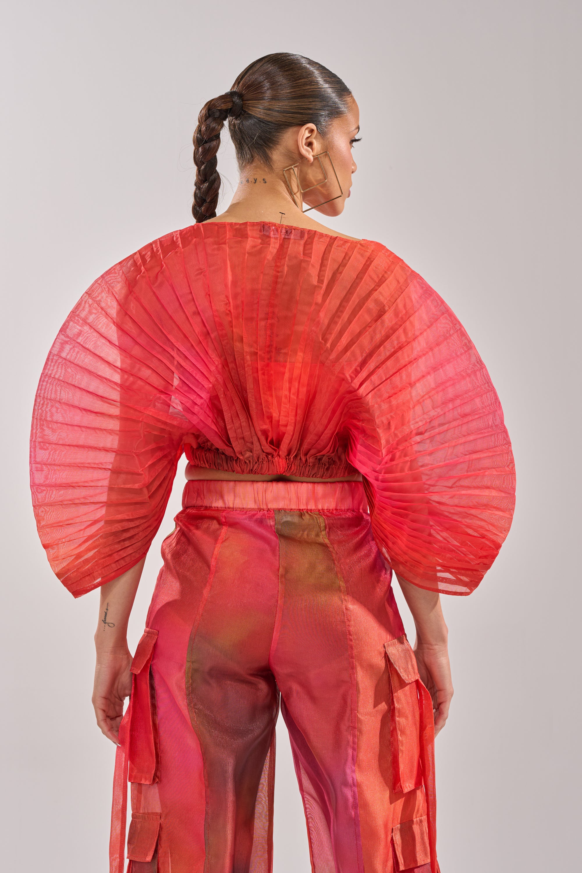 A woman with braided hair and large hoop earrings stands with her back to the camera, wearing the STAINED RED CROPPED BLOUSE featuring sheer chiffon fabric, wide pleated sleeves, and matching layered red pants. 3