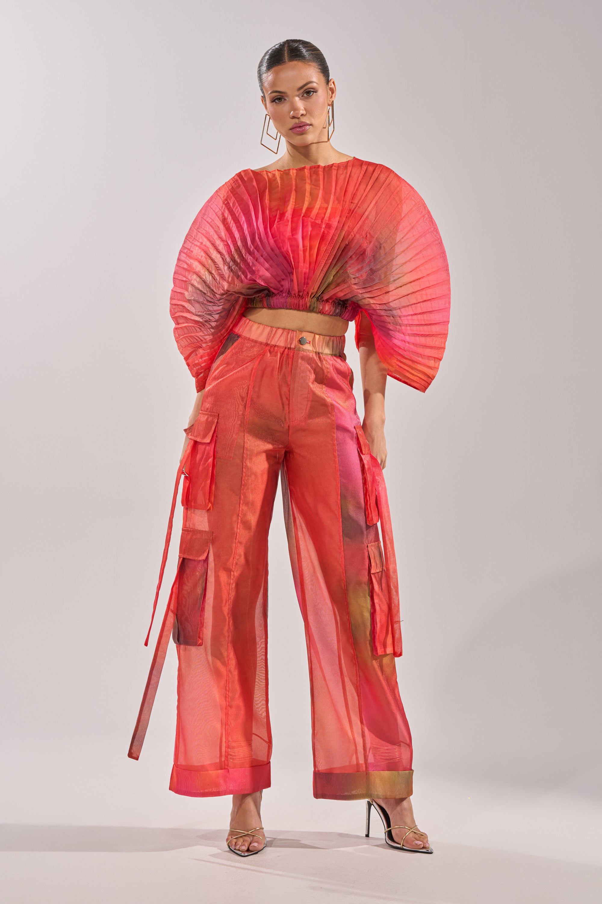 A woman models the STAINED RED CROPPED BLOUSE, a sheer chiffon top, styled with wide-leg pants, hoop earrings, and clear high heels against a plain light background. 4