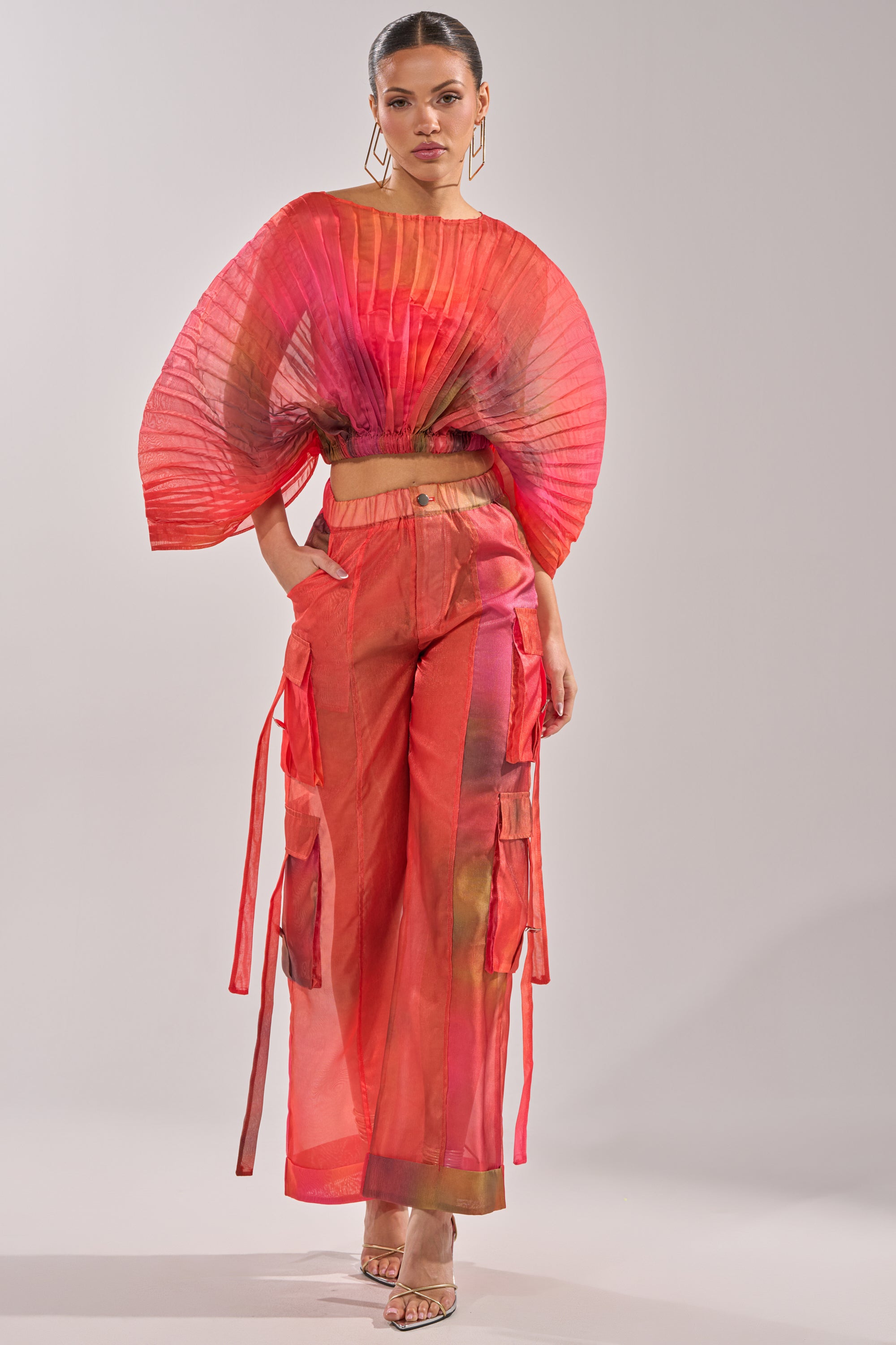 A woman stands confidently against a light background, wearing the STAINED RED CROPPED BLOUSE in bold, sheer chiffon with a pleated fan design, wide-legged pants, large geometric earrings, and strappy heels. 5