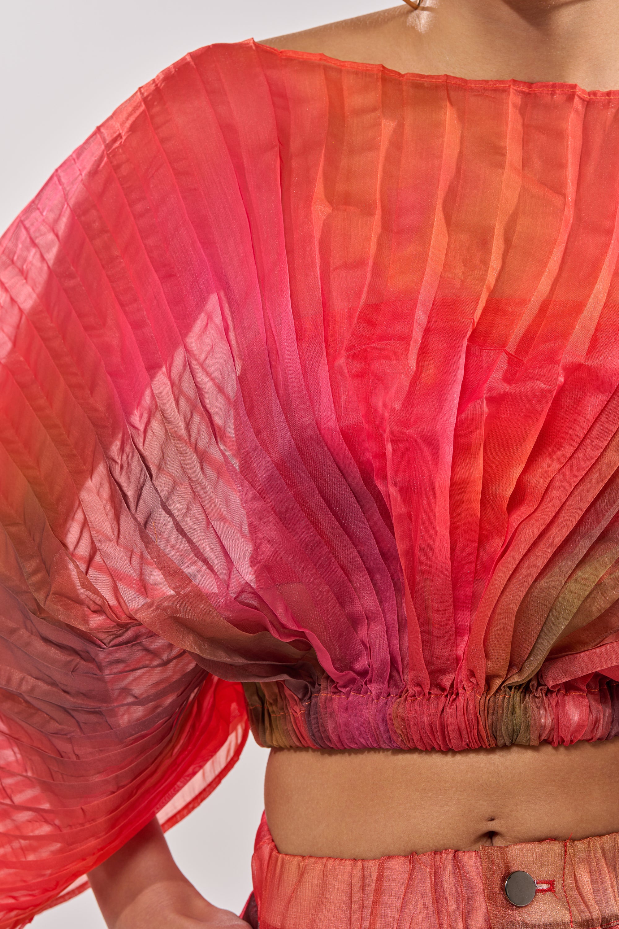 A close-up of a person wearing the STAINED RED CROPPED BLOUSE, a vibrant, watercolor orange-red top with batwing sleeves, pleats, and a gathered waist, paired with matching high-waisted pants for an eye-catching stained effect. 7