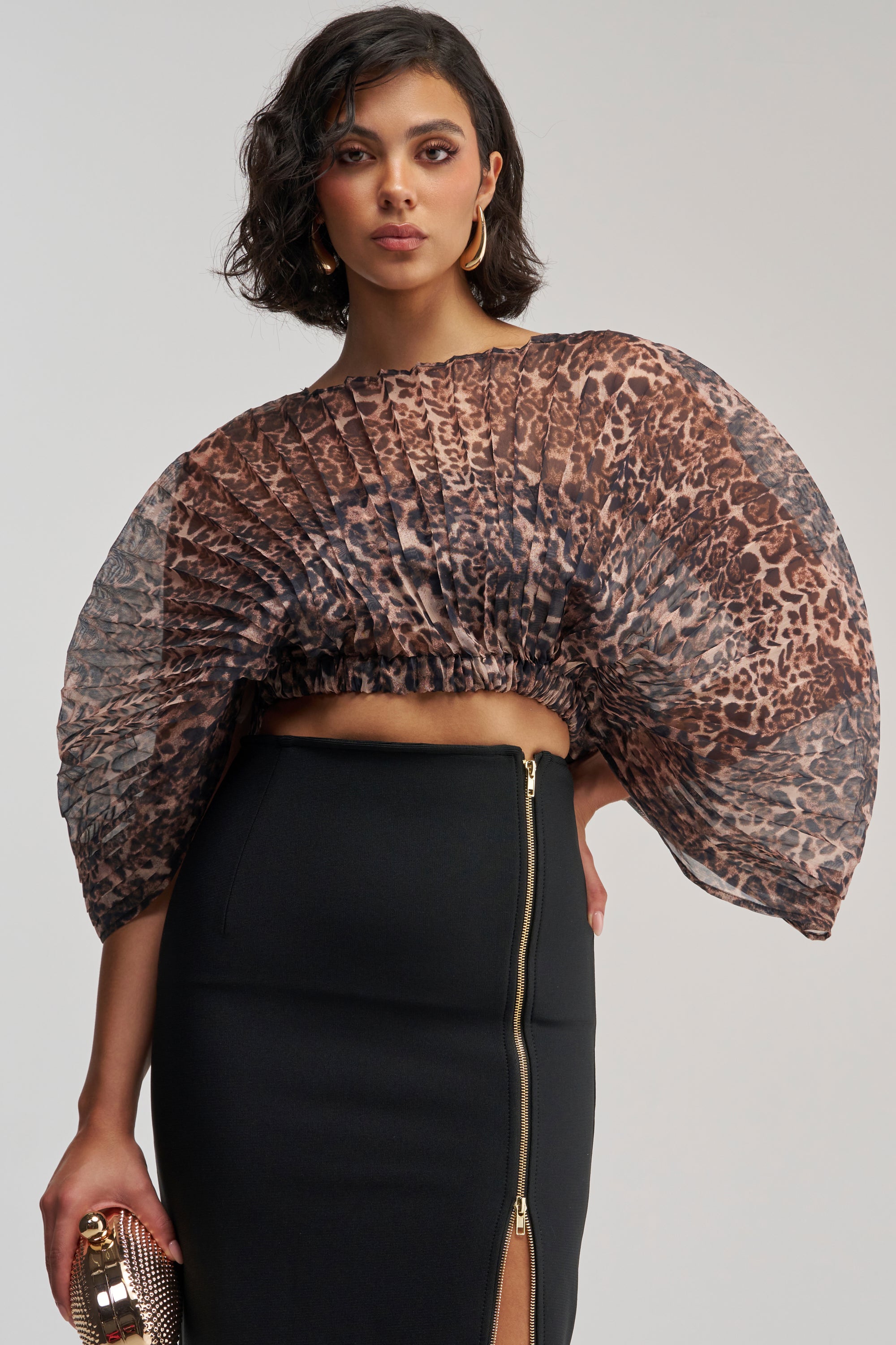 A woman wearing the UNTAMED INSTINCT CHIFFON TOP—a sheer, pleated fan blouse in cheetah print with voluminous sleeves—paired with a high-waisted black skirt featuring a front zipper and slit, holding a small clutch and standing against a plain background.