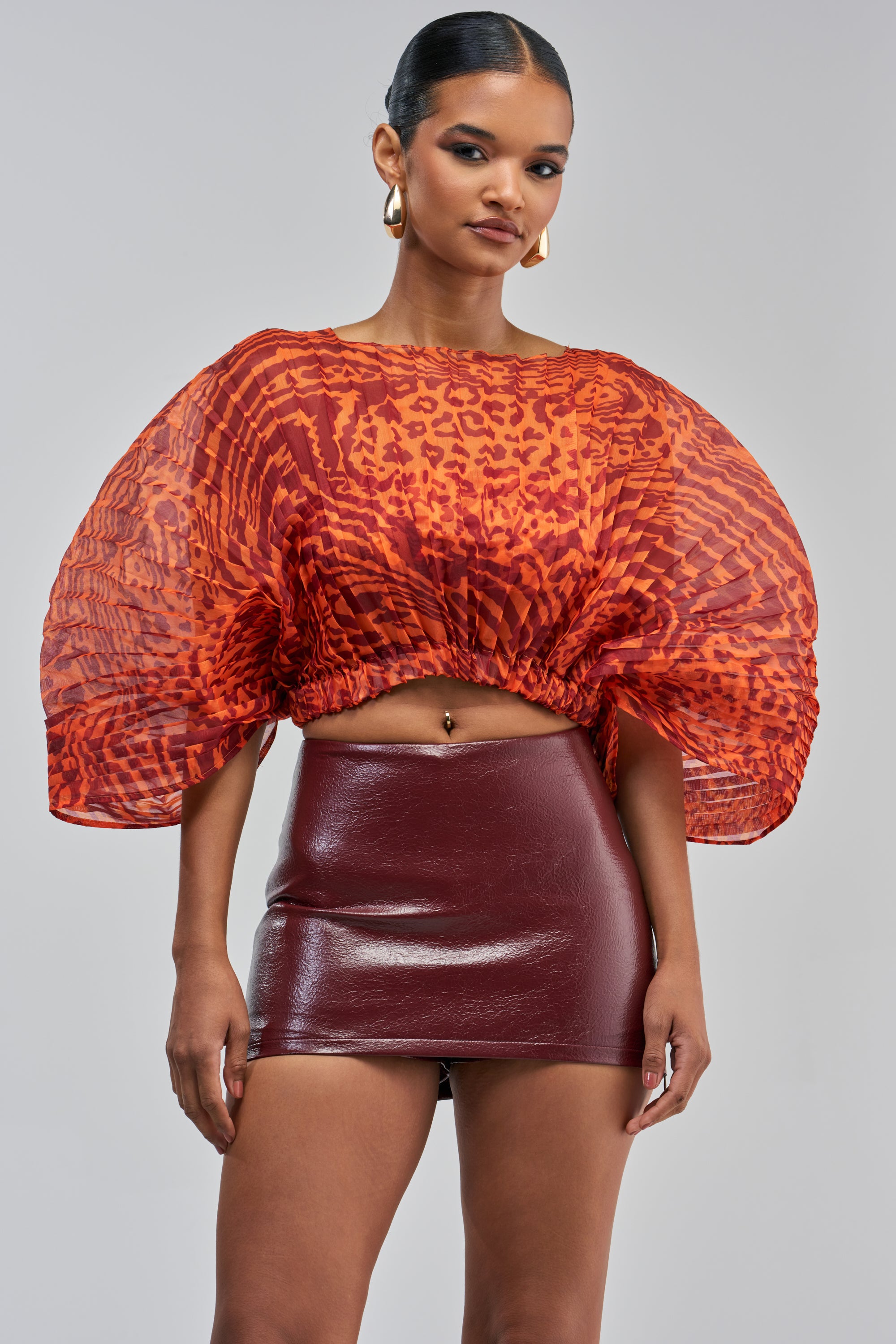 A woman exudes effortless chic in the WILDFIRE ENERGY CHIFFON TOP—a sheer, orange animal print blouse with puffed sleeves—paired with a shiny maroon mini skirt, standing confidently against a plain gray background and redefining modern resortwear.