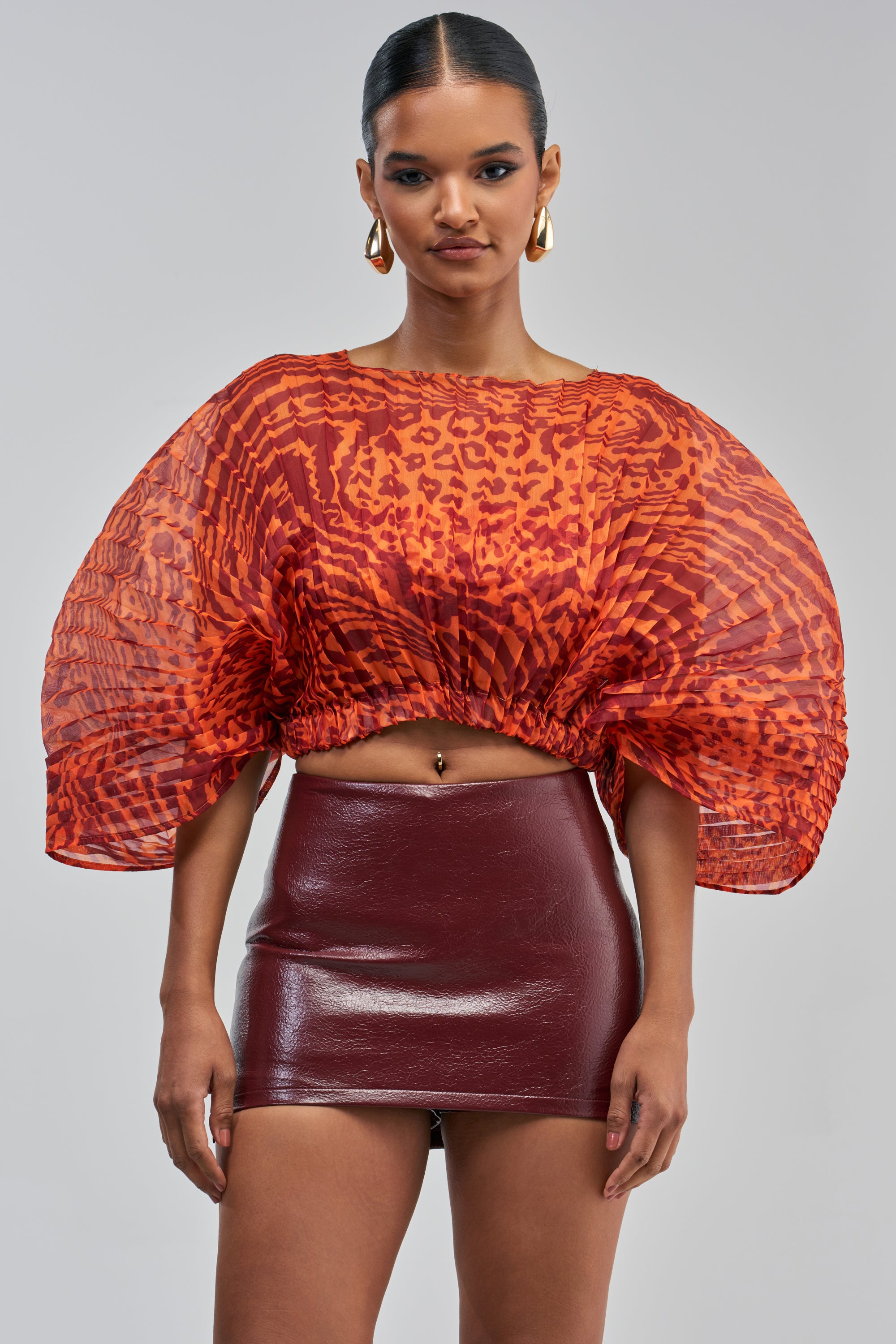 A woman wears the WILDFIRE ENERGY CHIFFON TOP, a sheer orange animal print blouse with large pleated sleeves, styled with a shiny burgundy mini skirt. She accessorizes with gold hoop earrings and has her hair pulled back.