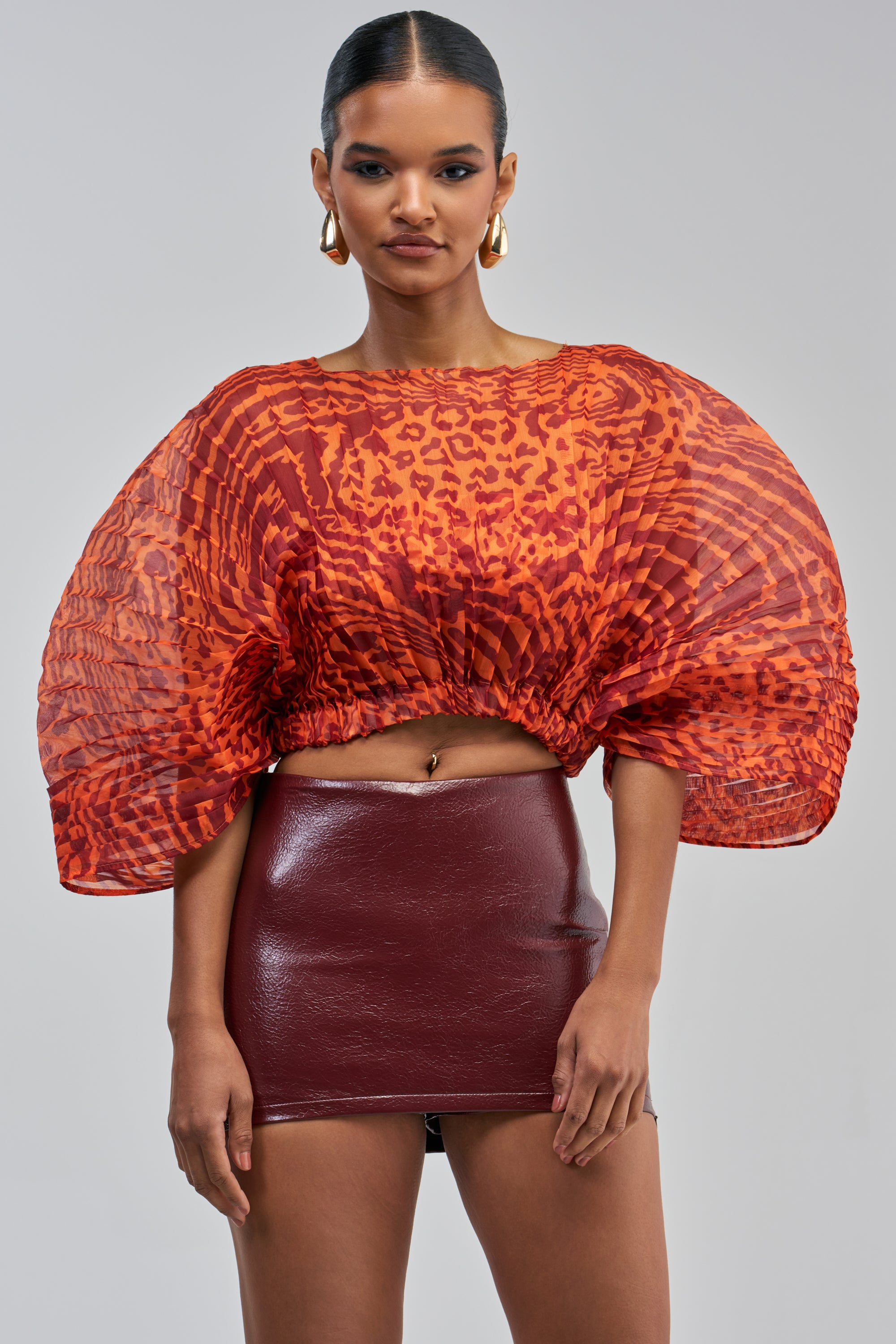 A woman stands against a plain gray background, looking at the camera, dressed in the WILDFIRE ENERGY CHIFFON TOP—a red and orange animal print blouse with puffed sleeves—and a shiny burgundy mini skirt.