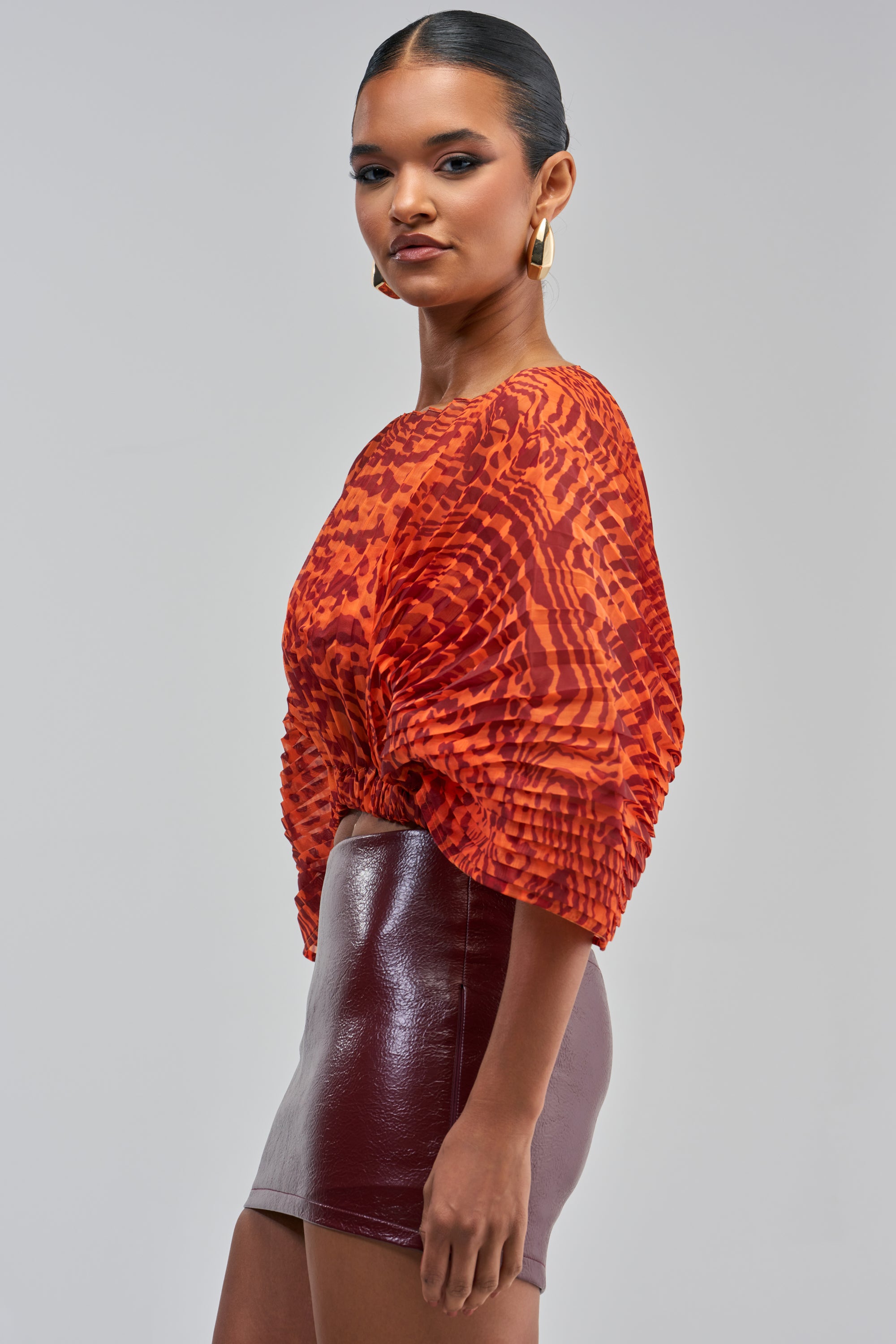 A woman showcases the WILDFIRE ENERGY CHIFFON TOP—a vibrant red patterned blouse with voluminous sleeves—paired with a shiny dark red mini skirt, posing against a plain light background for bold resortwear inspiration.