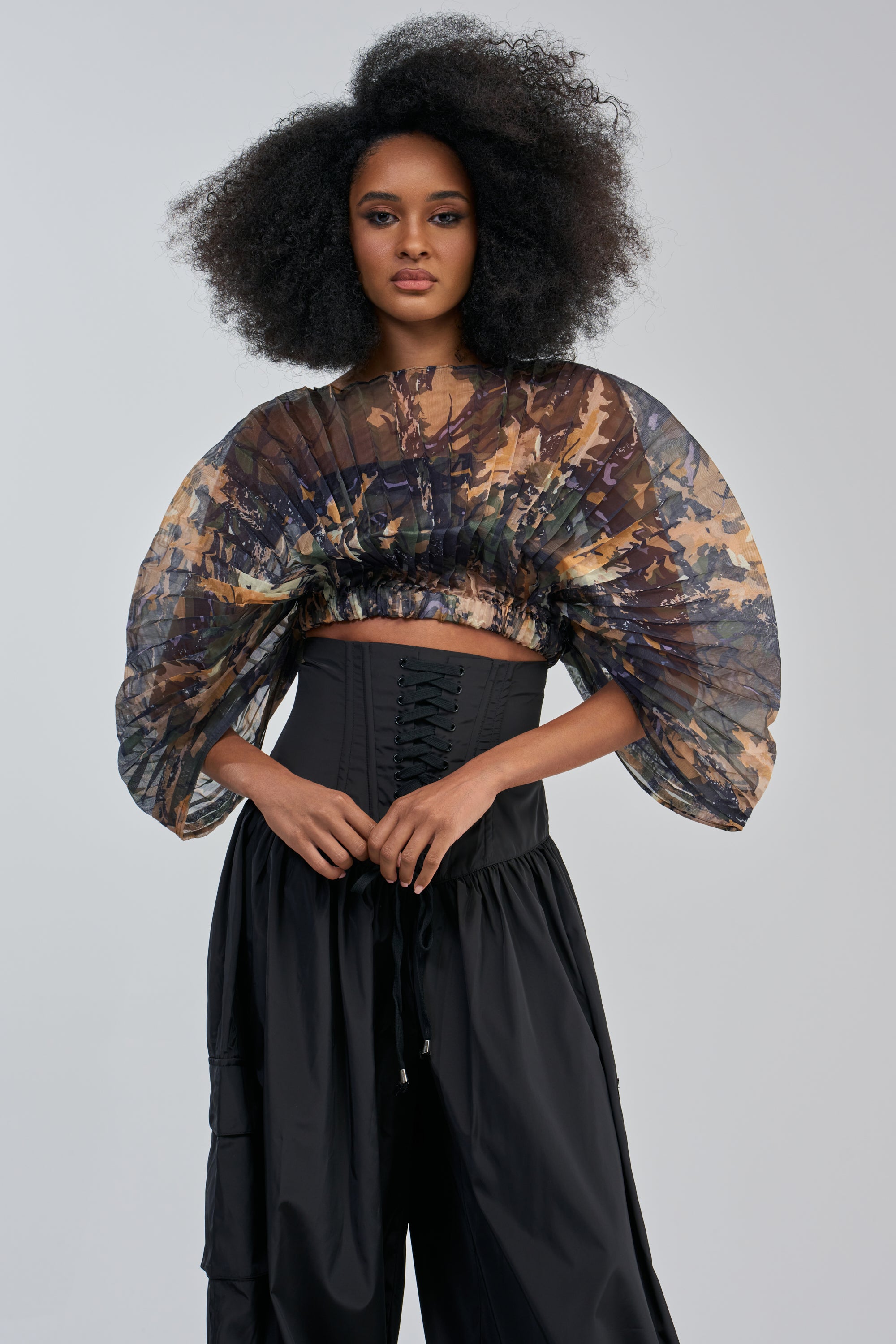 A woman with voluminous curly hair wears the MIDNIGHT MIRAGE CAMO TOP, featuring wide, flowing pleated sleeves, styled with a fitted black corset and loose black pants, standing against a light gray background.
