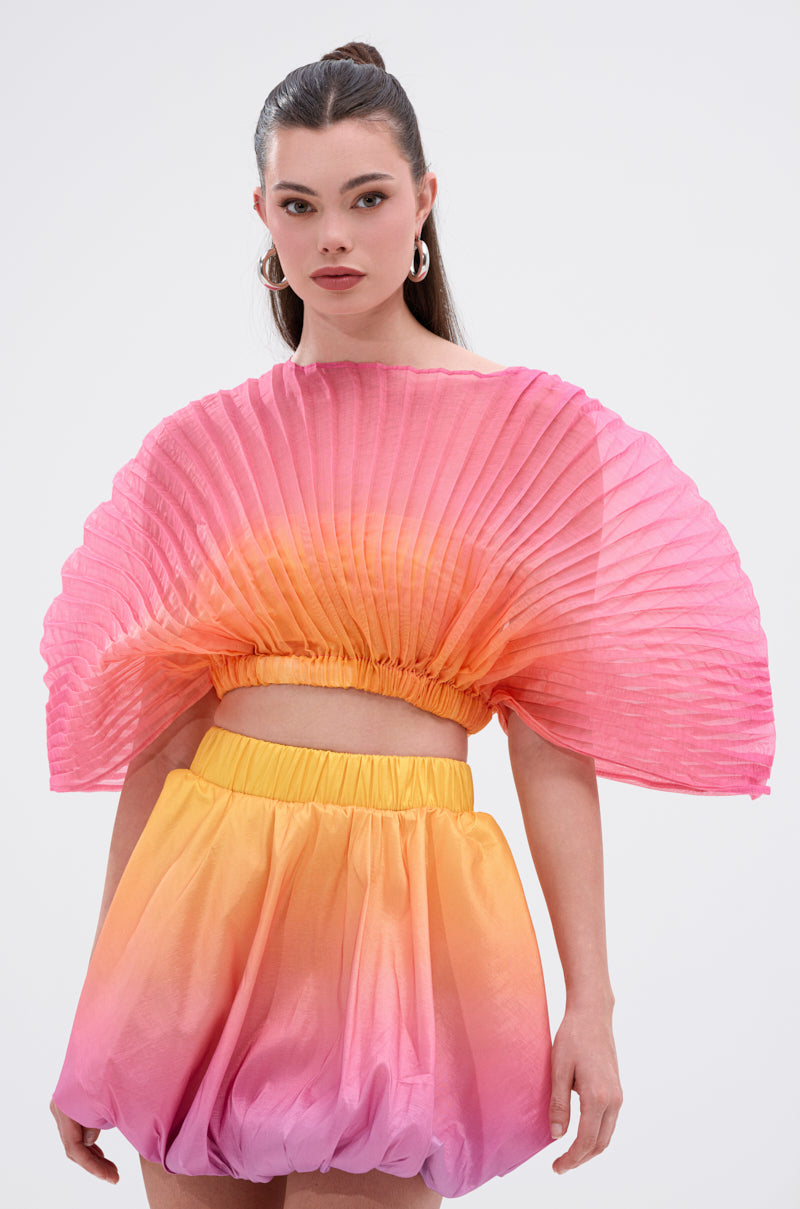 A woman wears the SHELBY OMBRE BLOUSE, a pleated, fan-shaped pink and orange crop top with a matching bubble skirt in a pink, orange, and yellow gradient, standing against a plain white background.