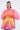 A woman wears the SHELBY OMBRE BLOUSE, a pleated, fan-shaped pink and orange crop top with a matching bubble skirt in a pink, orange, and yellow gradient, standing against a plain white background.