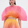 A woman wears the SHELBY OMBRE BLOUSE, a pleated, fan-shaped pink and orange crop top with a matching bubble skirt in a pink, orange, and yellow gradient, standing against a plain white background.