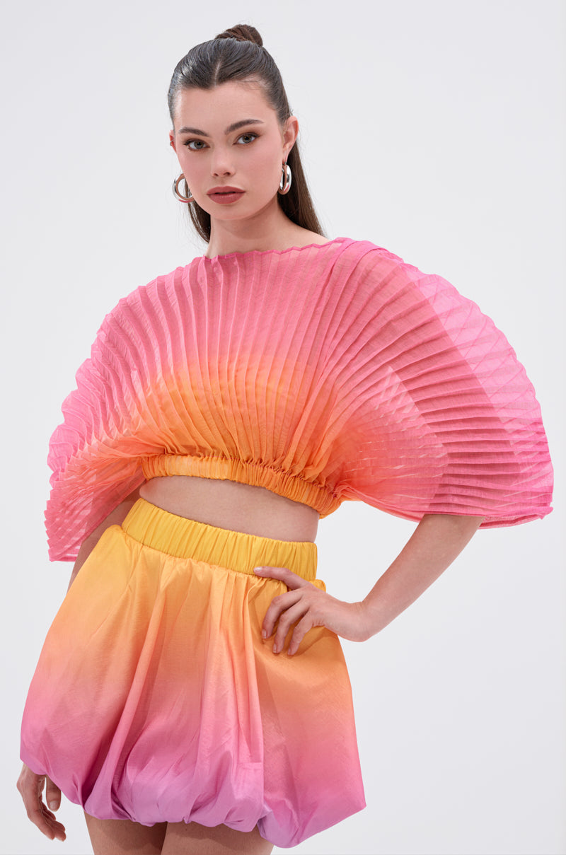 A woman wears the SHELBY OMBRE BLOUSE, featuring vibrant pink, orange, and yellow hues with dramatic fan sleeves, paired with matching bubble shorts. Her hair is tied back and she accessorizes with hoop earrings.