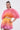A woman wears the SHELBY OMBRE BLOUSE, featuring vibrant pink, orange, and yellow hues with dramatic fan sleeves, paired with matching bubble shorts. Her hair is tied back and she accessorizes with hoop earrings.