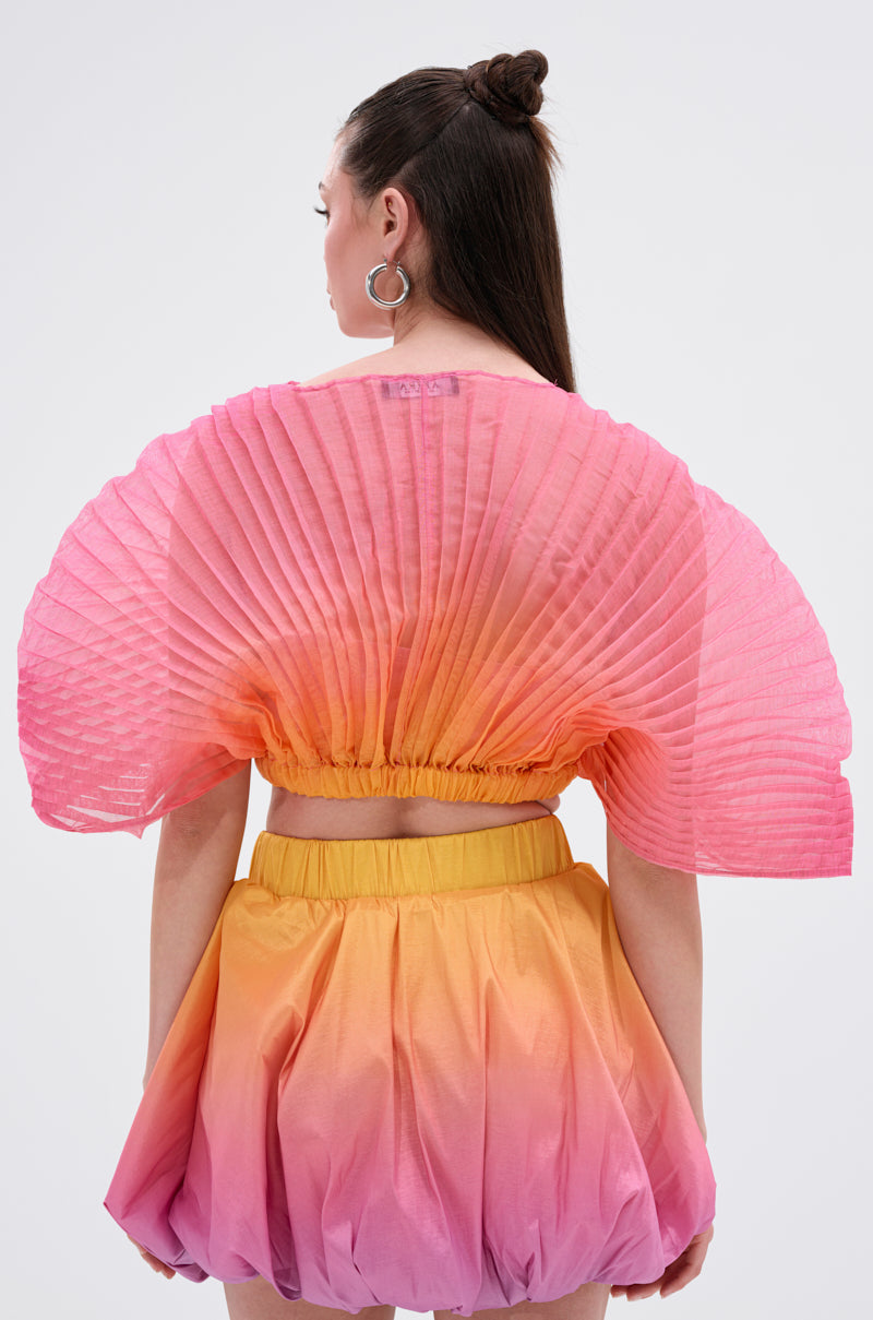 A person with long dark hair in a half-up bun is seen from the back, wearing the SHELBY OMBRE BLOUSE, which features a vibrant ombre design and pleated, wing-like top, paired with a puffy skirt against a plain light background.