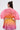 A person with long dark hair in a half-up bun is seen from the back, wearing the SHELBY OMBRE BLOUSE, which features a vibrant ombre design and pleated, wing-like top, paired with a puffy skirt against a plain light background.
