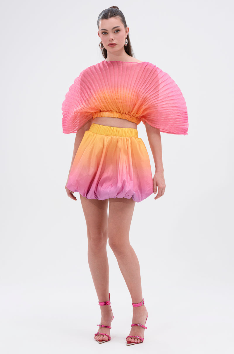 A woman poses against a white background wearing the SHELBY OMBRE BLOUSE, a pleated pink-and-orange crop top with matching puffed shorts in stretchy mesh, styled with pink high-heeled sandals.