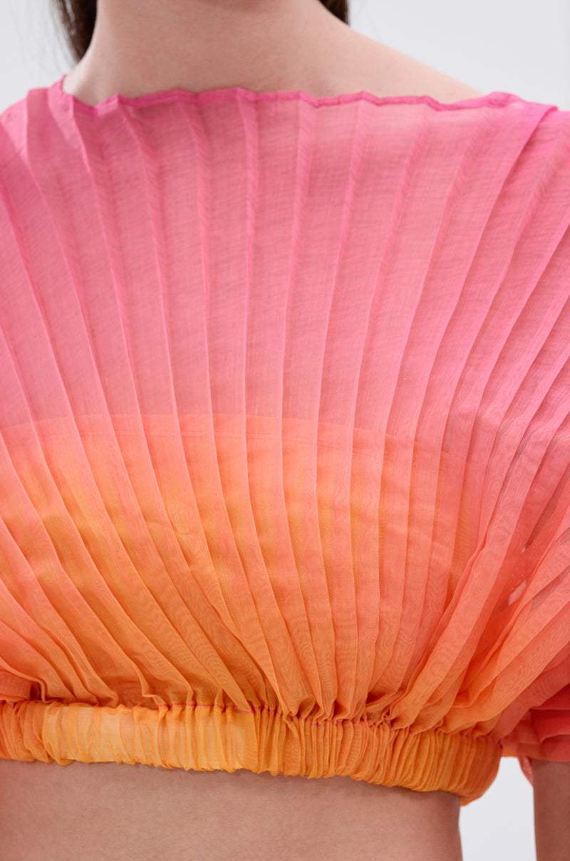 A close-up of someone in the SHELBY OMBRE BLOUSE, showcasing vibrant pleated mesh with a pink-to-orange-and-yellow gradient, gathered at the hem for a soft ombre effect.
