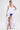 A woman models the TOO COOL HIGH LOW TOP, a dramatic white halter-style high-low top layered over denim shorts. She accessorizes with a textured white clutch and white heels, her sleek hair pulled back, against a plain white background.
