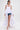 A woman wears the TOO COOL HIGH LOW TOP in white over blue denim shorts, paired with a textured white clutch, white heels, statement earrings, and a sleek ponytail.