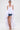 A woman poses against a plain white background wearing the TOO COOL HIGH LOW TOP, a sleeveless halter-style high-low top with voluminous ruffles cascading over denim shorts and silver strappy sandals.