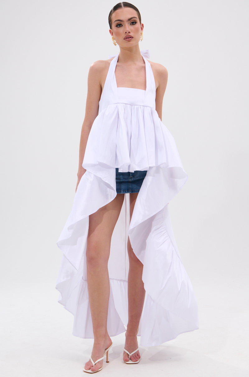 A woman poses against a white background wearing the TOO COOL HIGH LOW TOP—an eye-catching halter-style piece with cascading ruffles and a flowing train over denim shorts, styled with white heeled sandals and gold earrings. 3