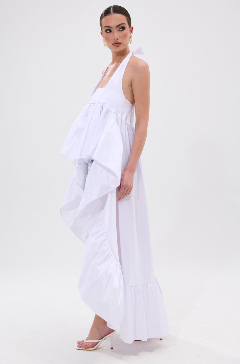 A woman wears the TOO COOL HIGH LOW TOP, a white halter-style piece with voluminous ruffles and an asymmetrical high-low hem, paired with white heeled sandals against a plain white background. 4