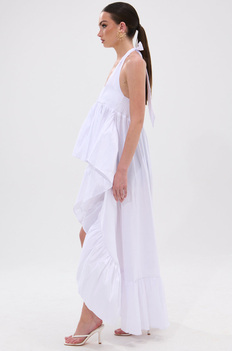 A woman stands in profile wearing the TOO COOL HIGH LOW TOP—an asymmetrical, white halter-style top with ruffles—paired with large gold earrings, white heeled sandals, and a ribbon in her hair against a plain white background. 5