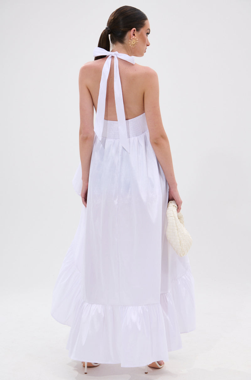 A woman stands facing away in the TOO COOL HIGH LOW TOP, a sleeveless white halter-style top with ruffles and a ruffled hem. She holds a white woven clutch, wears large earrings, has her hair in a low ponytail, against a plain white background. 6
